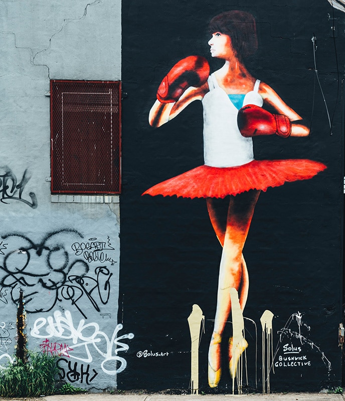 Image of ballerina wearing a tutu, with a white top, red skirt wearing red boxing gloves next to a wall with graffiti.