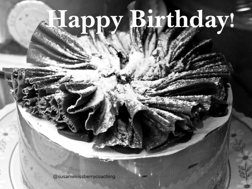Black and white image of a fancy cake topped with rolled chocolate shavings. with the word Happy Birthday in white at the top,.